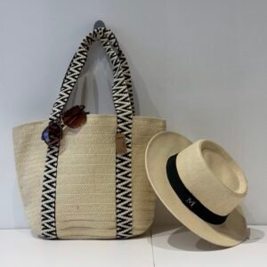 Bolso tote tejido color natural con asas en patrón blanco y negro colgado en pared blanca, acompañado de una visera negra con lazo elegante. Accesorios de moda playa con estilo sofisticado y minimalista.